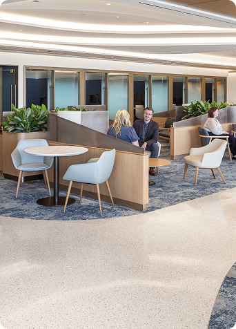 A spacious, bright lounge with upholstered chairs, tables, partitioned seating areas, green plants, and several people seated or walking.