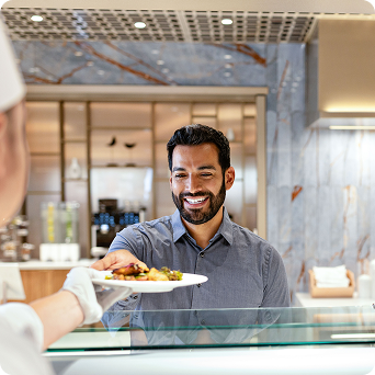 A smiling man with dark hair and a beard, wearing a gray shirt, reaches to accept a plate of food from a staff member across a glass food-service counter.