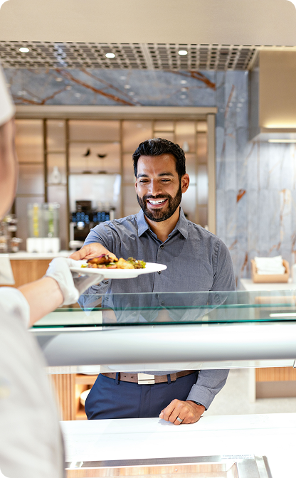 A smiling man with dark hair and a beard, wearing a gray shirt, reaches to accept a plate of food from a staff member across a glass food-service counter.