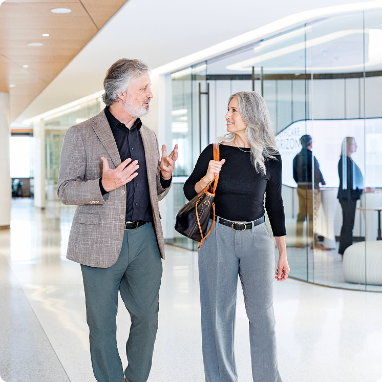 A gray-haired man in a checked blazer gestures while walking and talking with a gray-haired woman wearing a black top and gray trousers and carrying a shoulder bag. They look at each other in a bright corridor lined with glass-walled rooms.