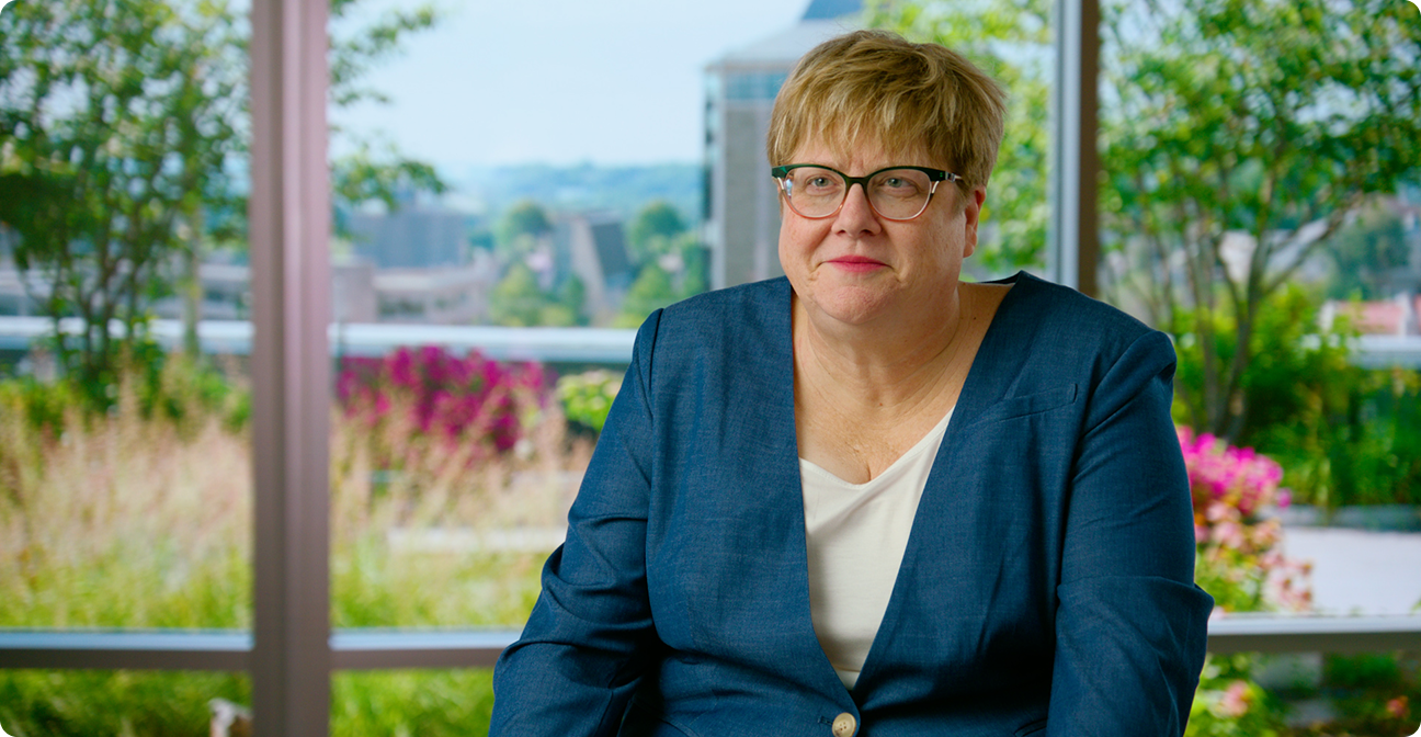A woman with short blond hair and glasses, wearing a teal blazer over a white top, sits in front of large windows overlooking green trees, pink flowers, and distant buildings.