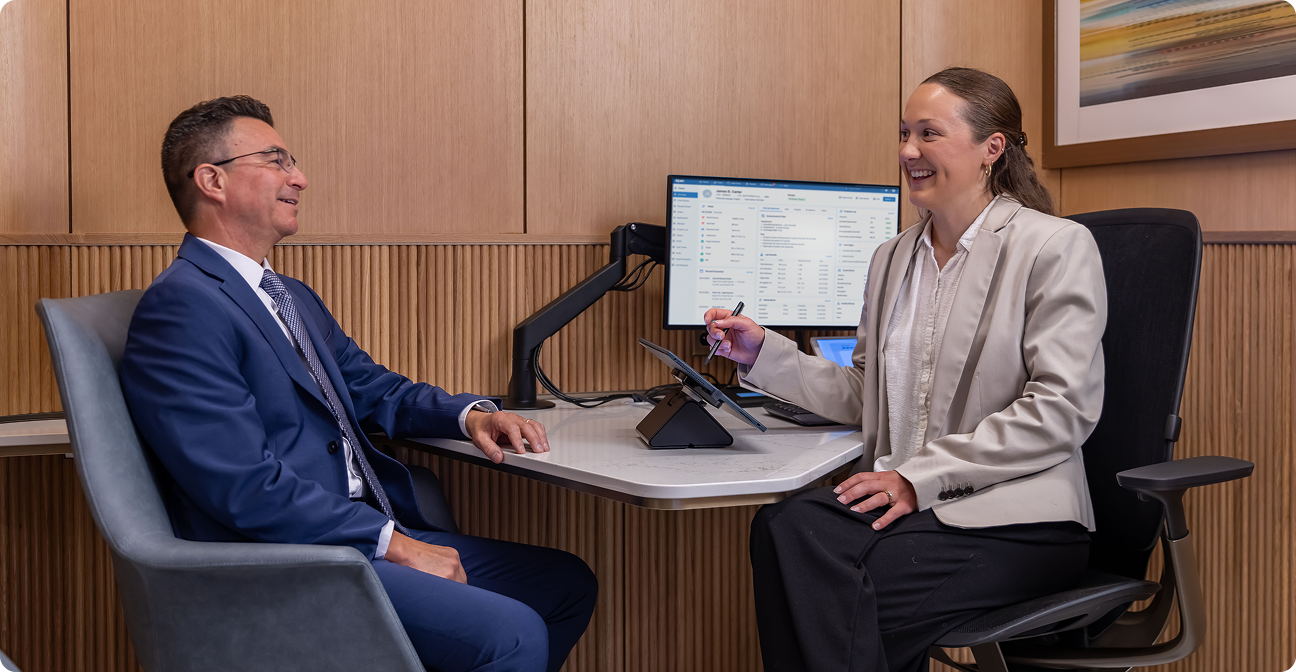A woman in a light gray blazer holds a pen and smiles while speaking with a man wearing glasses and a blue suit across a desk in a wood-paneled office. A tablet and computer monitor sit on the desk.