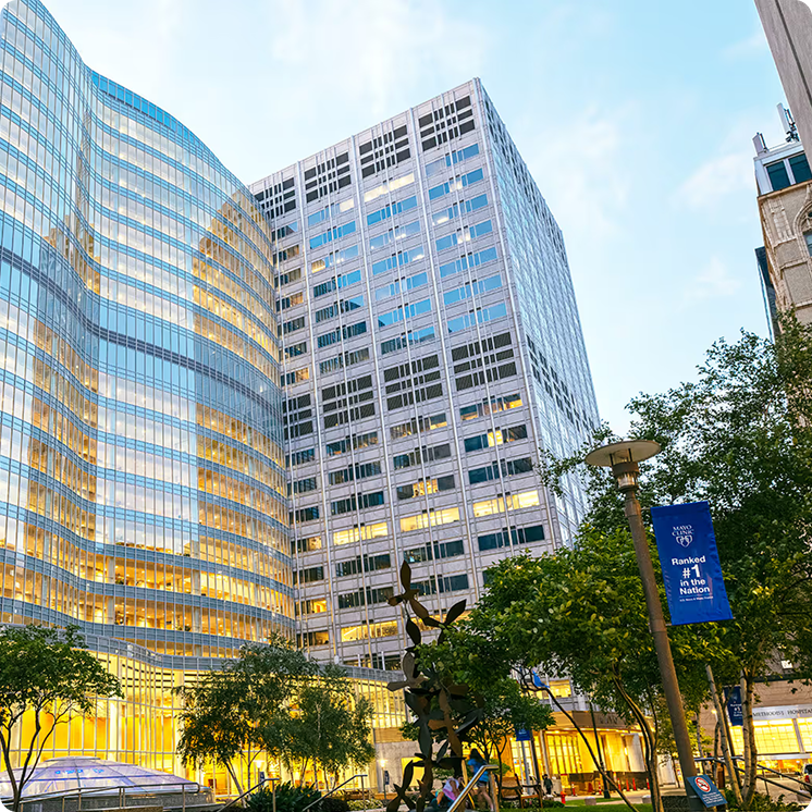 Exterior view looking up at the Mayo Clinic's curved glass tower flanked by stone-clad office buildings, with banners on the street lamps reading 'Mayo Clinic — Ranked #1 in the Nation.