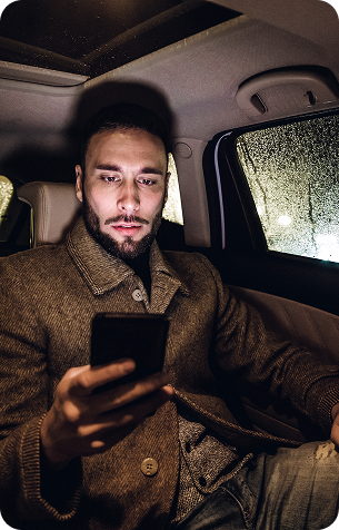 A bearded man in a tweed coat sits in the back seat of a car at night, looking at his phone, with rain streaking the side window.