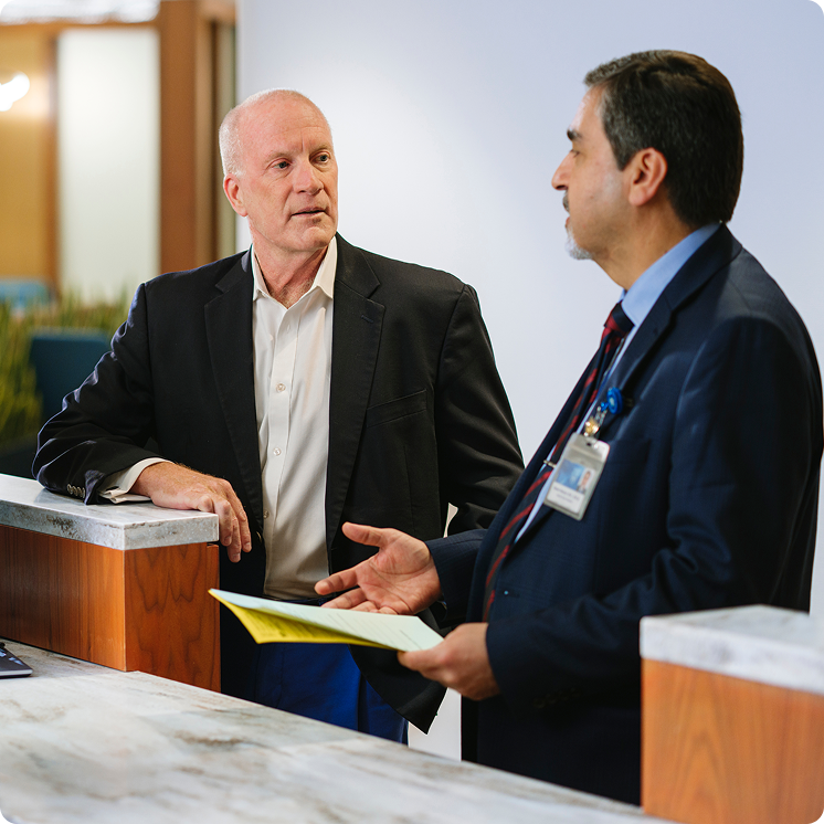 A bald man in a dark blazer leans on a marble counter, listening to a clinician in a navy suit and Mayo Clinic ID badge who gestures while holding a yellow form.
