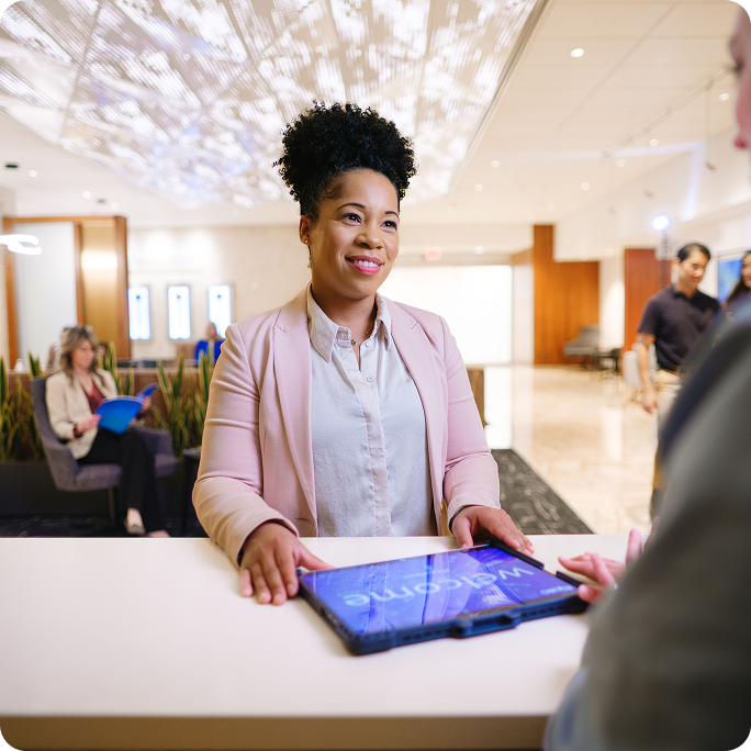 A smiling woman in a blush blazer stands behind a reception desk holding a tablet displaying 'Welcome,' speaking with a guest whose back is to the camera. A bright, modern lobby is visible behind her.
