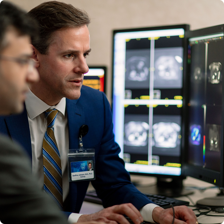 A radiologist in a blue suit and striped tie reviews medical imaging scans displayed across multiple monitors, while a colleague looks on from the foreground.