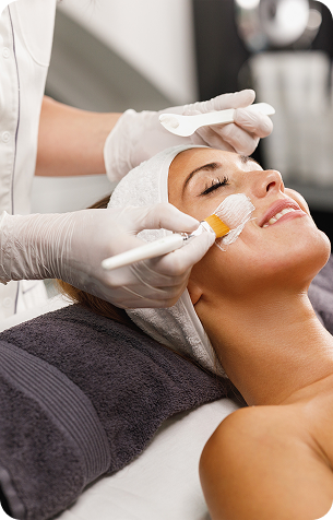 A woman with her eyes closed receives a facial treatment as a gloved esthetician applies product to her cheek with a brush.