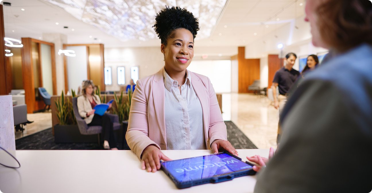 A smiling woman in a blush blazer stands behind a reception desk holding a tablet displaying 'Welcome,' speaking with a guest whose back is to the camera. A bright, modern lobby is visible behind her.