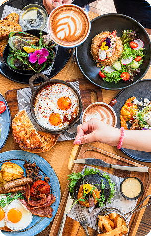 Overhead view of a wooden table set with plates of fried eggs, salads, toast, sausages, and two lattes with leaf-patterned foam art.