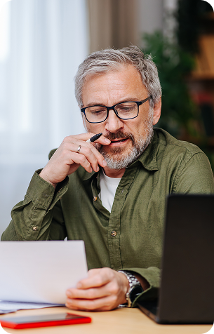 A grey-haired man in glasses and a green shirt rests his hand on his chin while reading a document at a laptop.