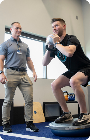 A trainer in a grey polo shirt stands beside a man performing a goblet squat with a kettlebell on a raised platform.