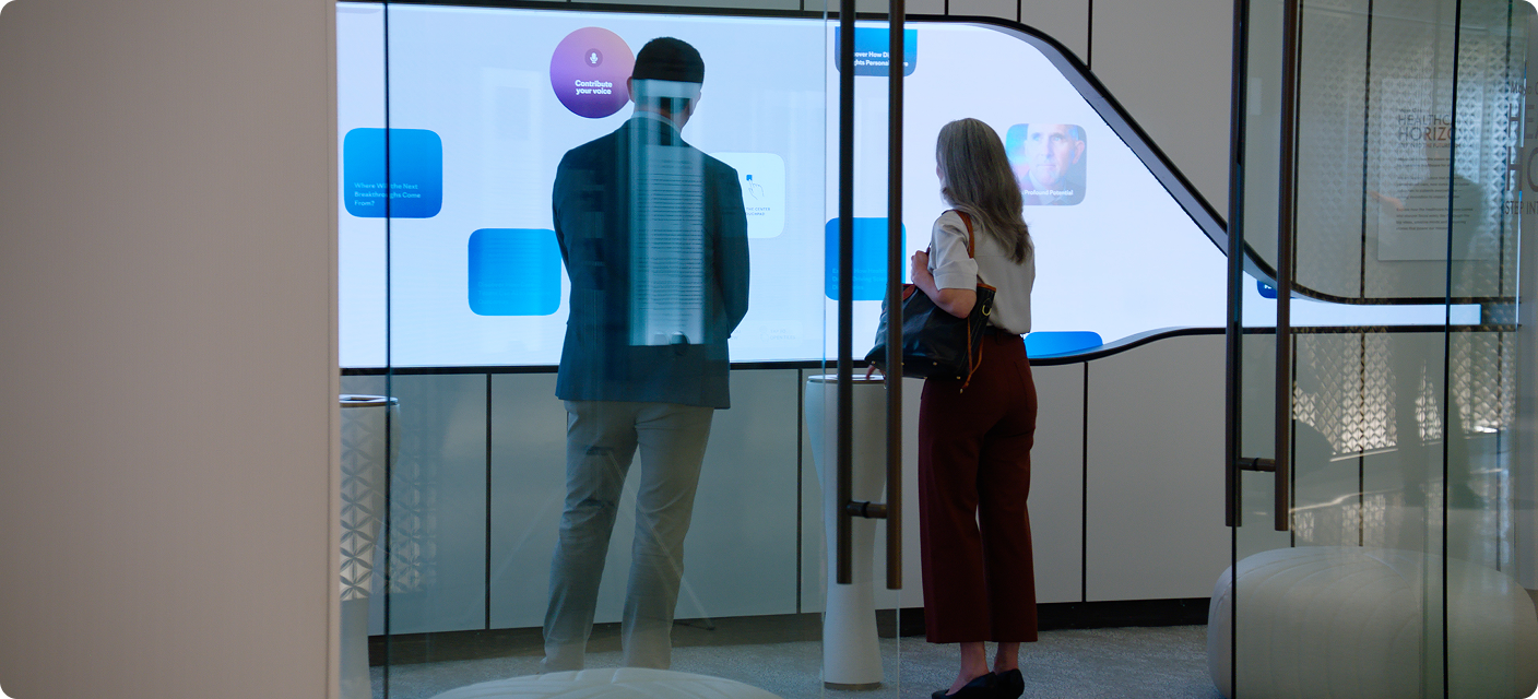 Two people, viewed from behind through glass doors, stand facing a large illuminated wall display with colorful information panels. A man wears a dark blazer and light trousers, and a woman wears a light top and rust-colored trousers and carries a shoulder bag.