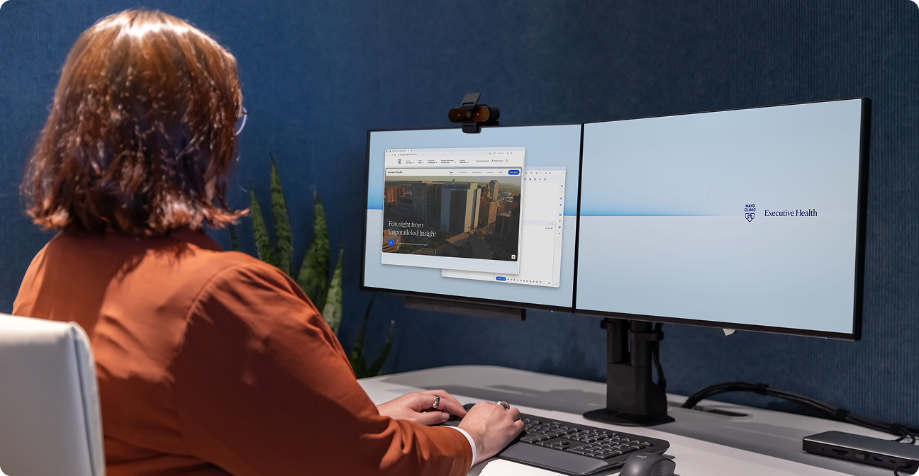 A person in an orange top works at a desk with a keyboard and two monitors in a blue-walled workspace.