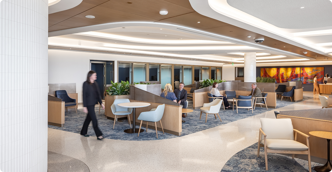 A spacious, bright lounge with upholstered chairs, tables, partitioned seating areas, green plants, and several people seated or walking.