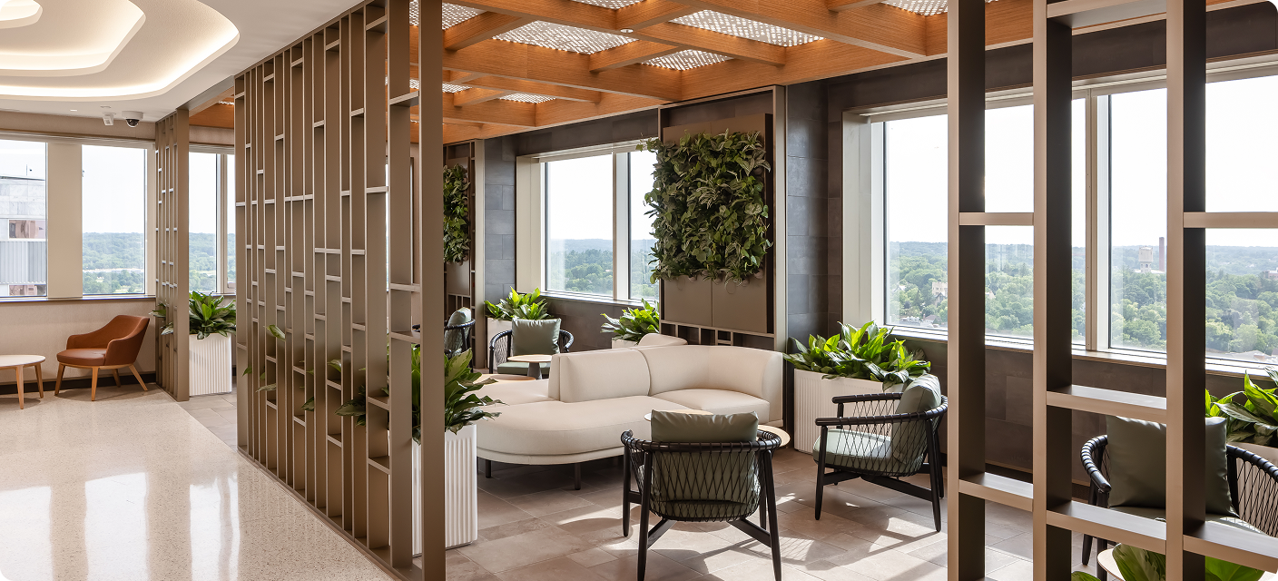 A quiet lounge seating area framed by open wood partitions, with a curved cream sofa, dark woven chairs, round tables, and abundant green plants. Large windows overlook trees and buildings.