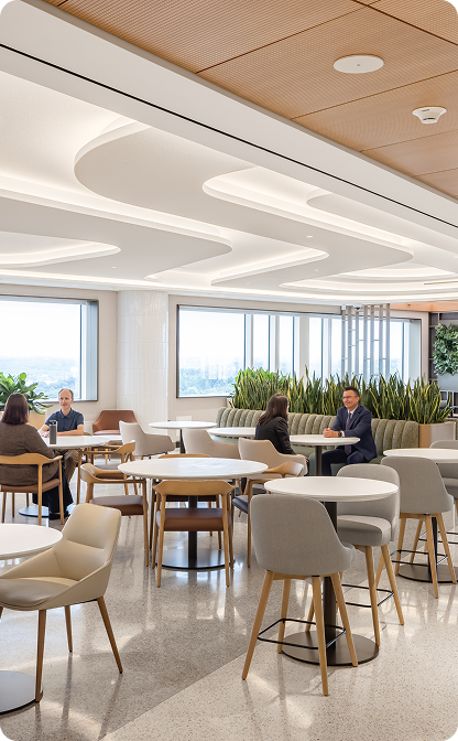 A bright, modern dining lounge with upholstered chairs, round tables, large windows, and tall green plants. Several people sit at tables beneath curved white ceiling features and wood paneling.