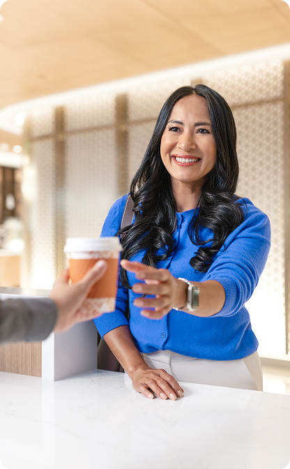 A smiling woman with long dark hair, wearing a blue cardigan, reaches to accept a takeaway cup offered by another person across a white counter.