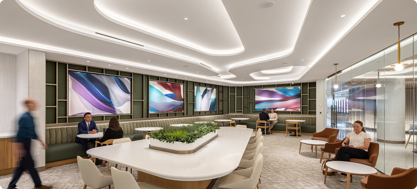 A spacious modern lounge with a long communal table surrounding a central planter, upholstered chairs, curved booth seating, and large abstract artworks on the walls. People sit and converse beneath sweeping illuminated ceiling features.