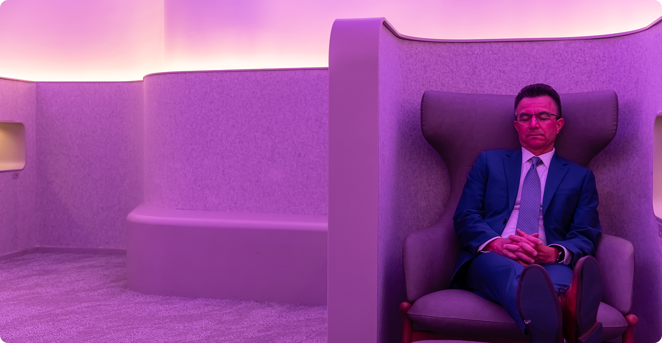 A man in a blue suit sits with his eyes closed and hands clasped in a high-backed chair within a private seating nook bathed in pink and purple light.