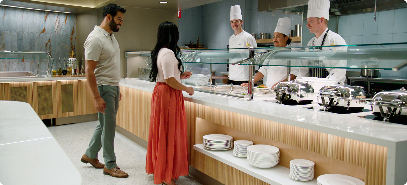 A woman in a white top and flowing orange skirt and a man in a light polo shirt stand at a food-service counter facing three chefs in white uniforms and tall hats. Covered serving dishes line the counter, with stacks of plates on a shelf below.