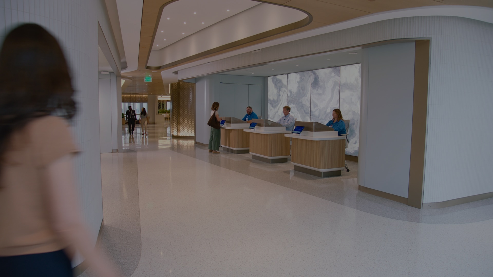 A woman approaches a reception area in a spacious, modern lobby. Three staff members stand behind curved wood-paneled desks with tablet screens, while another visitor speaks with one of them. Backlit stone panels and sweeping ceiling details frame the reception area.