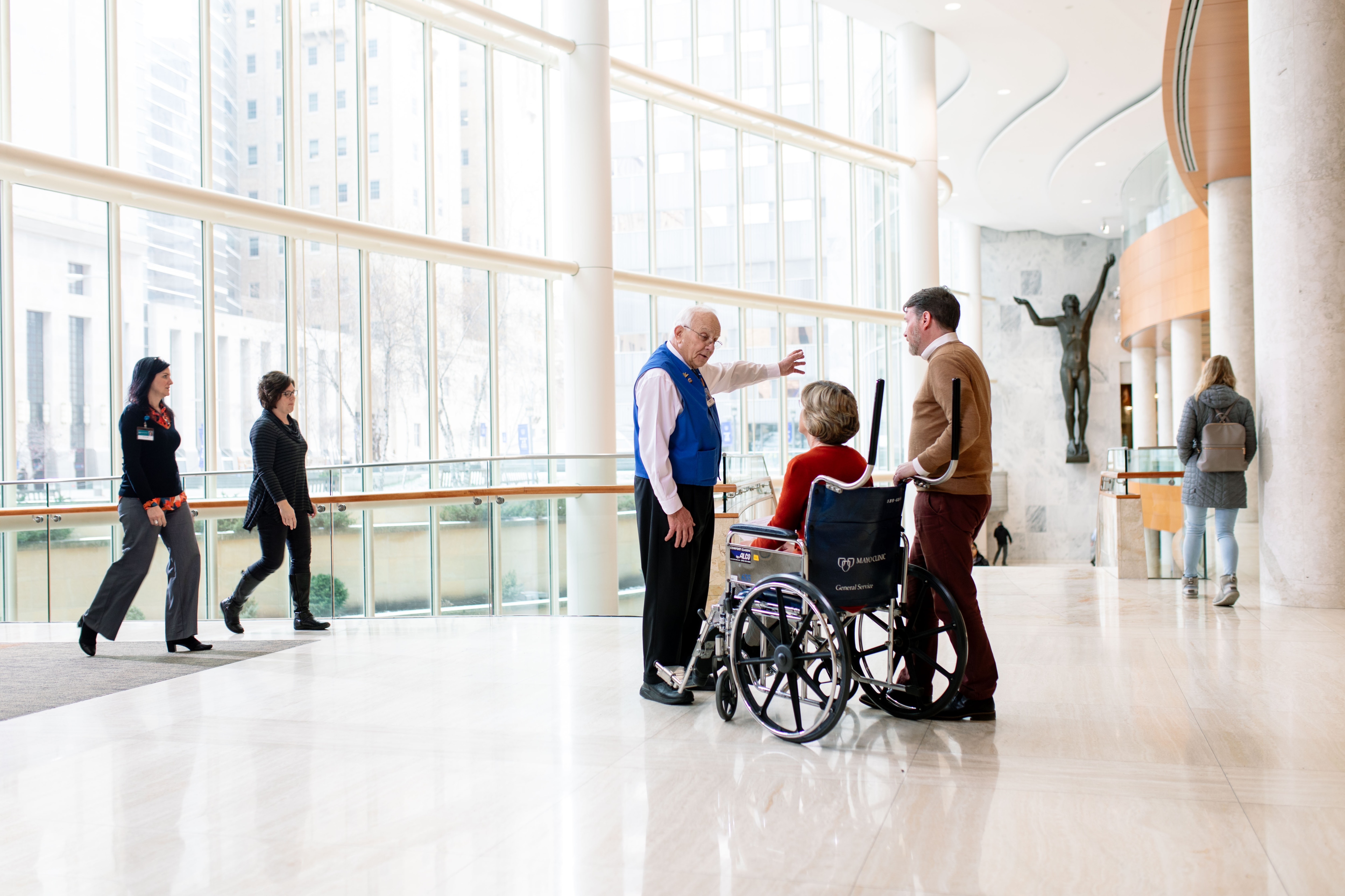 A person in a Mayo Clinic vest gives directions to a patient in a wheelchair and companion in a clinic atrium.