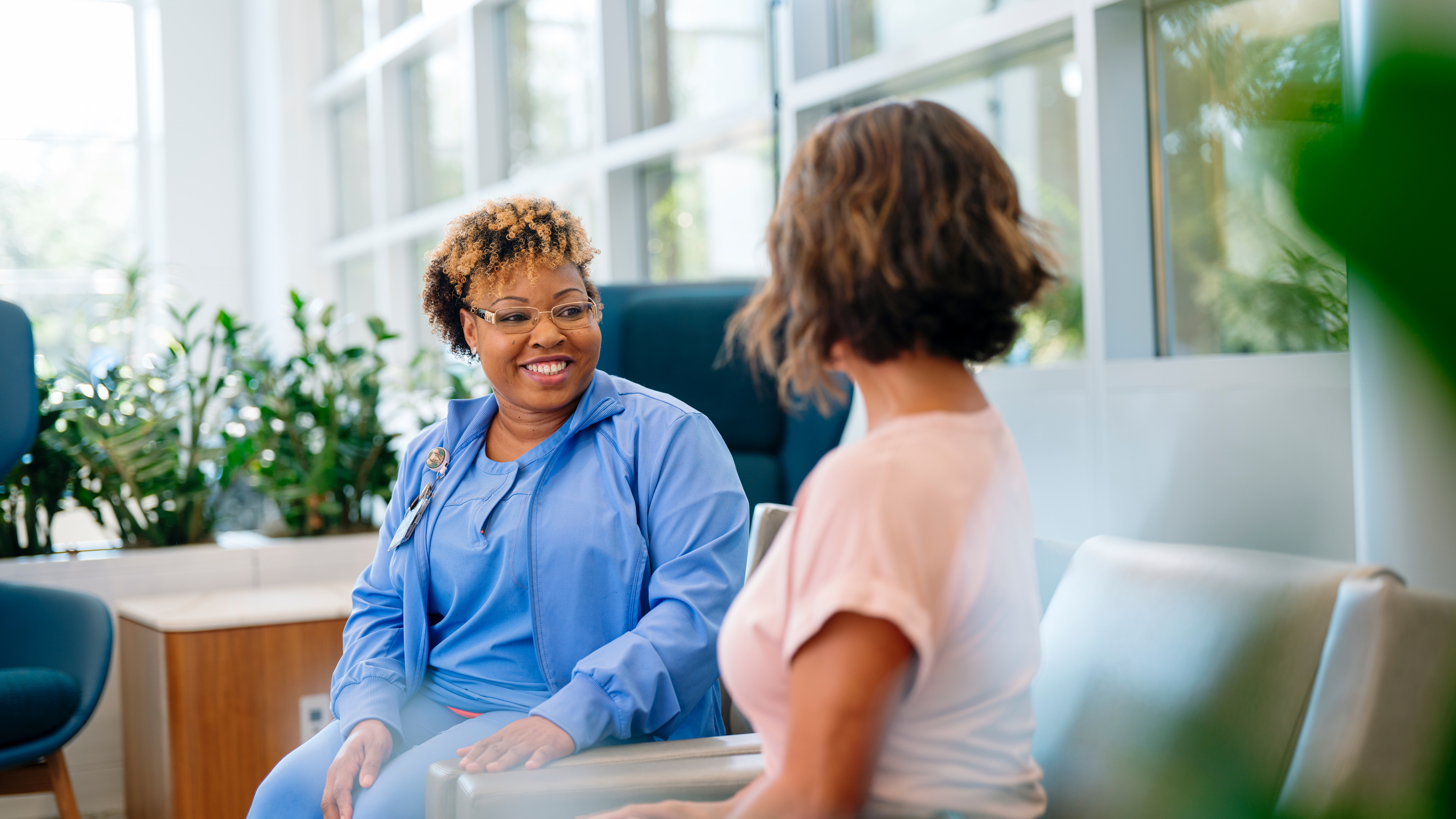 A nurse speaks with a patient in a lobby seating area.